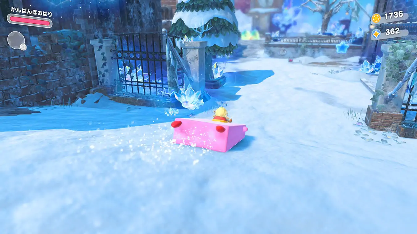 A gameplay screenshot showing Kirby using Mouthful Mode to stretch over a large, rectangular object. The player is sliding across a snowy courtyard path next to a stone building and an iron gate encrusted with blue ice crystals.