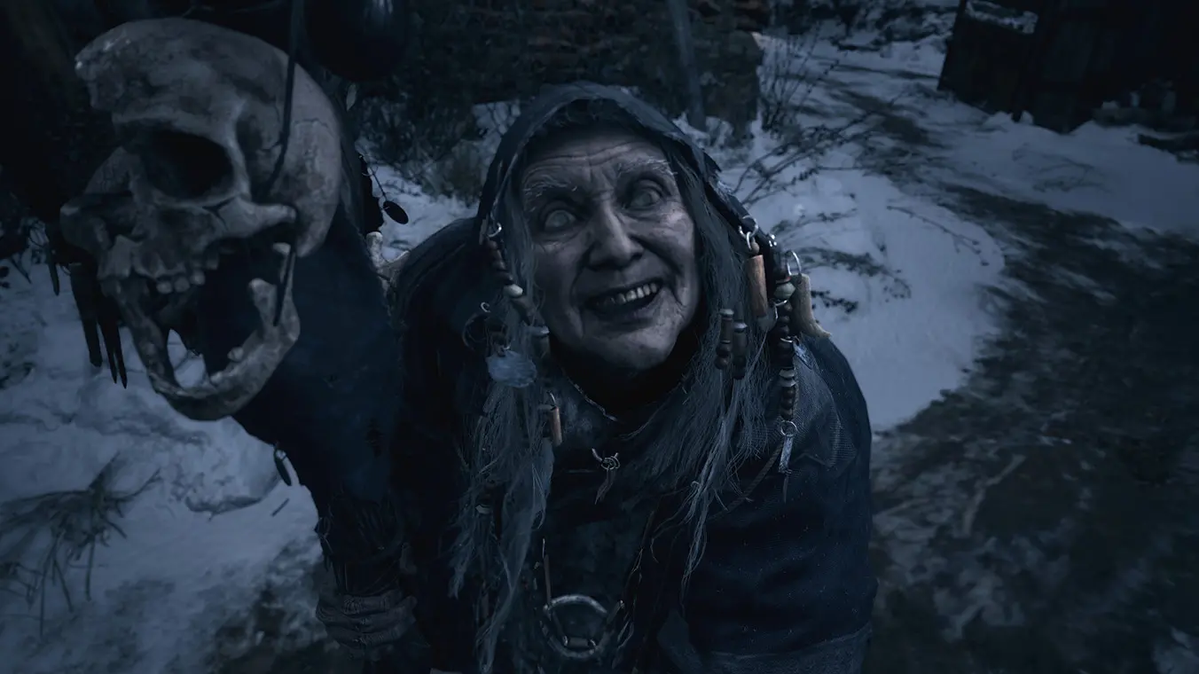 A close-up screenshot of an elderly, sinister-looking woman with gray hair, pale eyes, and a wide, unsettling grin. She is wearing a dark, hooded cloak adorned with trinkets and stands outdoors in a snowy environment next to a staff topped with a skull.